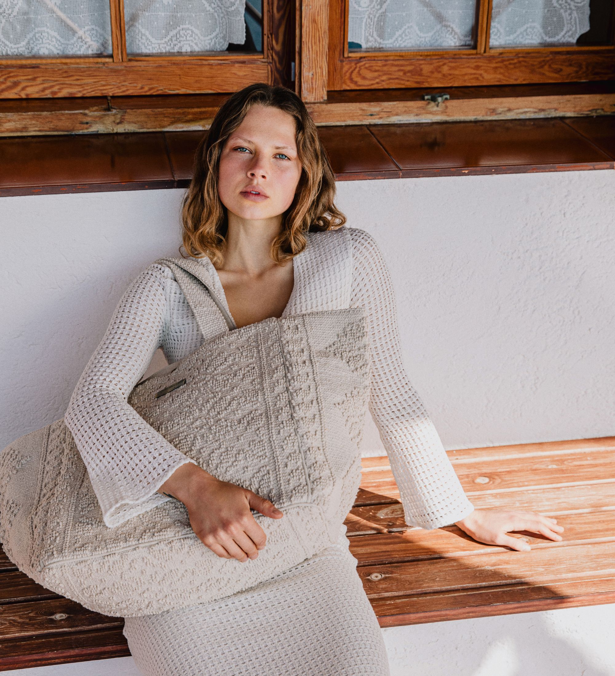 A woman in a textured white knit dress sits by a wooden window holding a large natural Sea You Soon woven tote in soft Mediterranean light.
