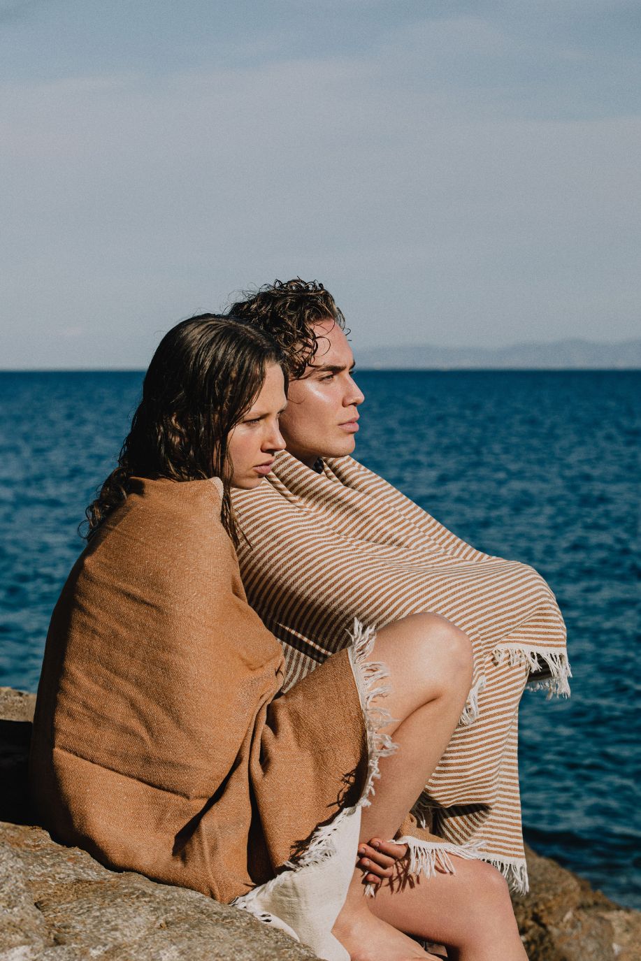 A man and woman sit wrapped in warm-toned striped Sea You Soon linen towels on coastal rocks, overlooking the calm Mediterranean sea.