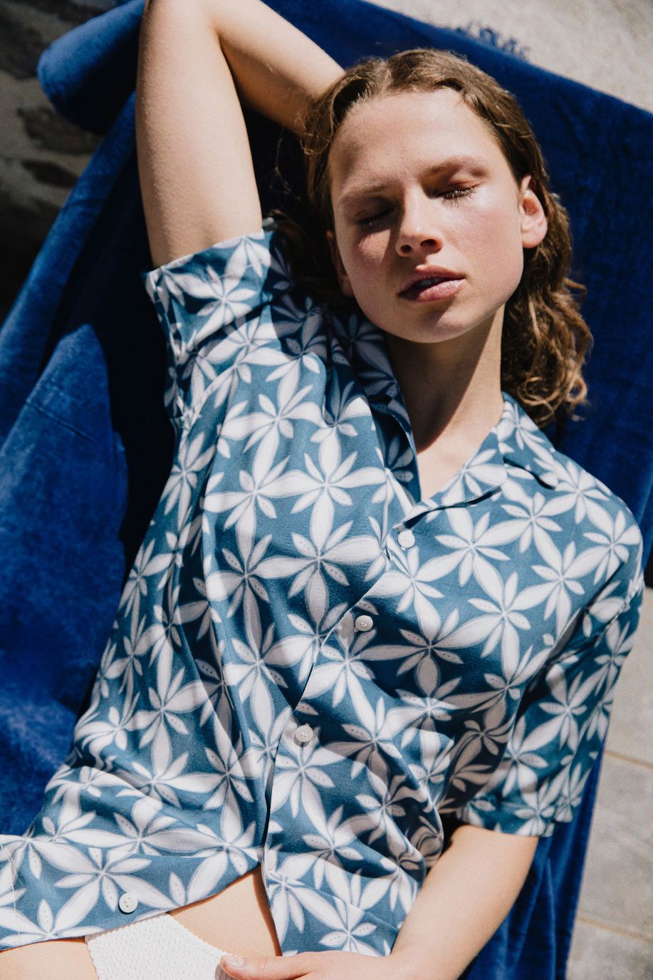 A woman relaxes in the sun on a blue towel wearing a blue patterned Sea You Soon short sleeve shirt with a white floral motif, captured in soft outdoor light.