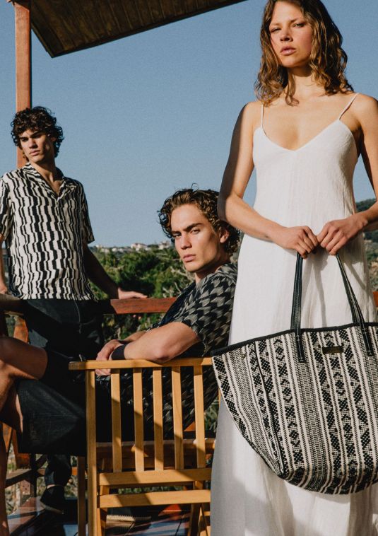 Three models in patterned summer shirts and a white dress stand on a sunlit terrace, with one holding a woven black-and-white Sea You Soon tote bag against a Mediterranean backdrop.