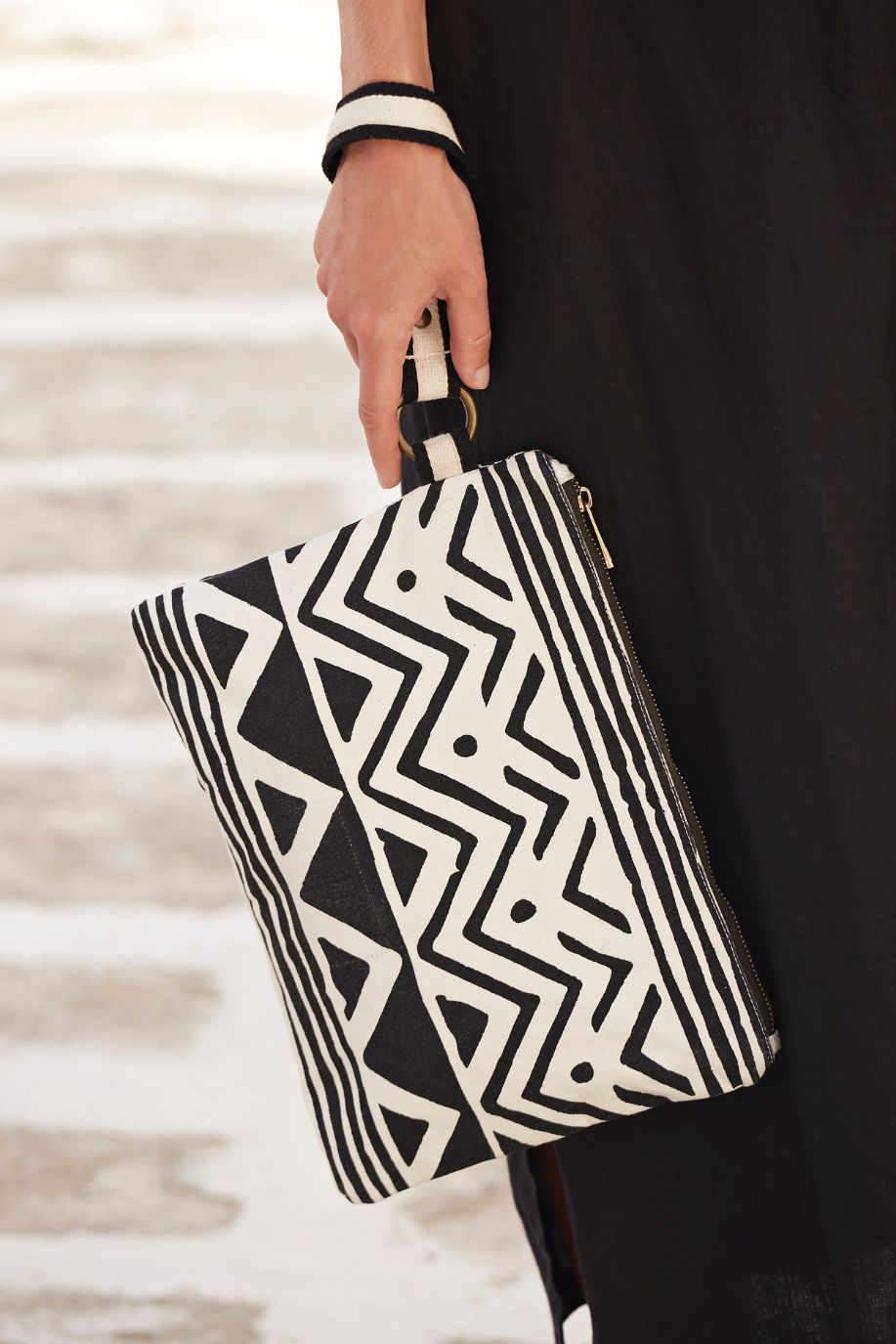 Close-up of a Sea You Soon geometric clutch bag in black and ecru canvas, held against a flowing black dress on a sunlit Mediterranean stone pathway.