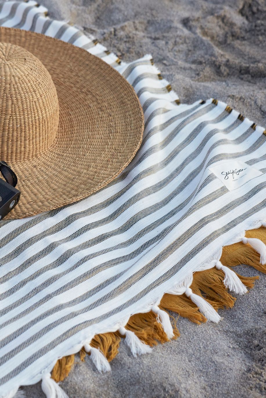 Straw hat and Sea You Soon striped beach towel with tassels laid on stone ground, styled with seaside accessories in soft Mediterranean light.