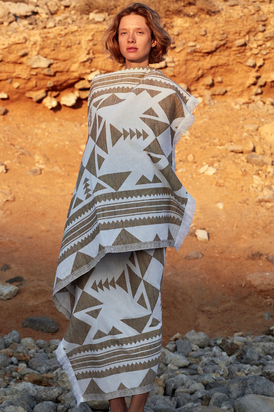 Woman wrapped in a Sea You Soon geometric cotton beach towel standing on rocky terrain in Antiparos, highlighting natural textures and earthy tones.
