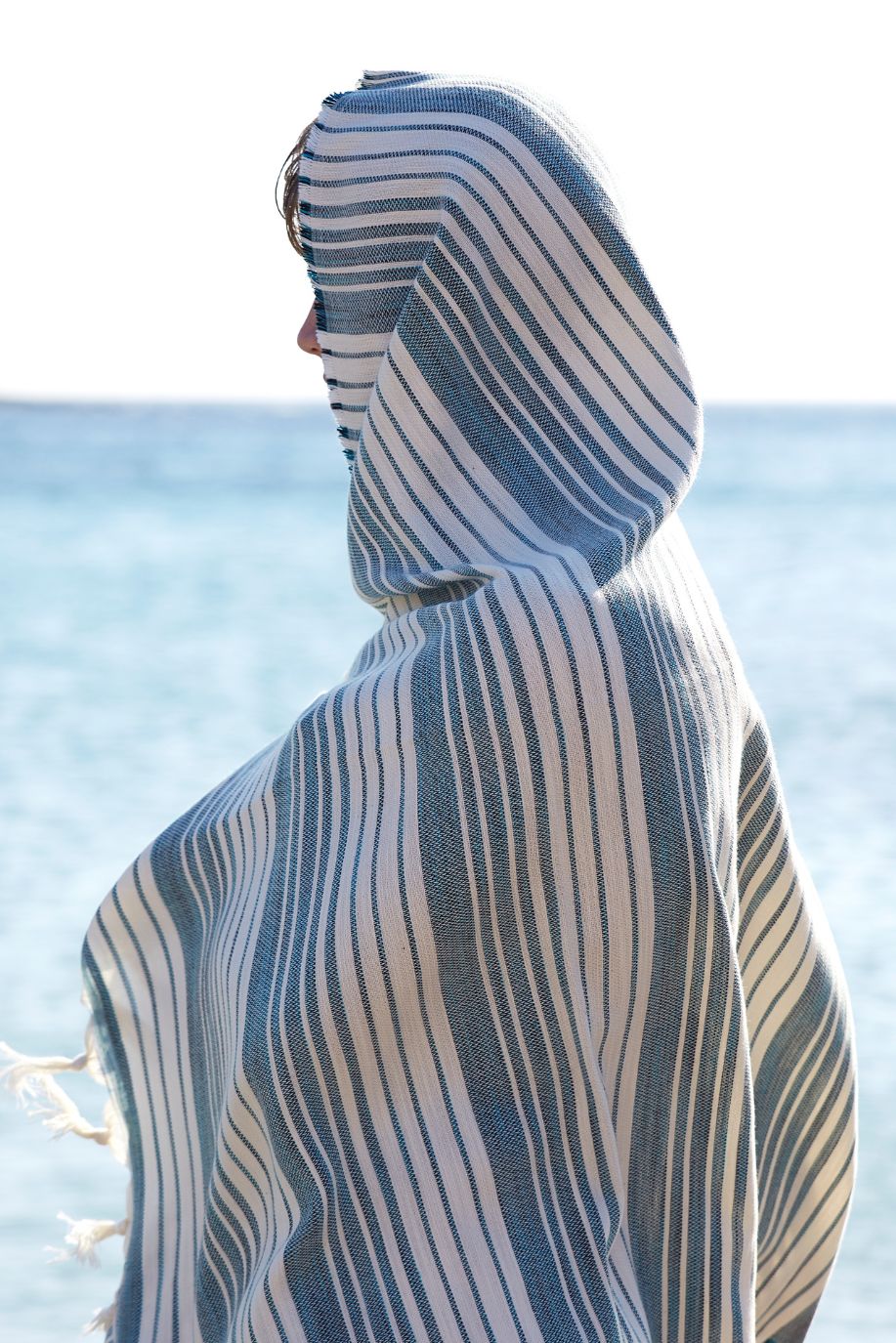 Figure wrapped in a Sea You Soon striped beach towel facing the sea, with calm Aegean waters and horizon lines framing the Antiparos setting.