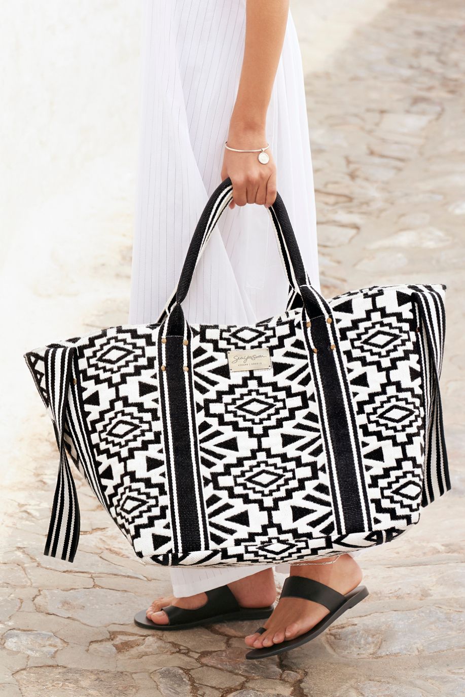 A woman in a white dress carries a black-and-white woven Sea You Soon tote bag while walking along a bright stone path beside a textured white wall.