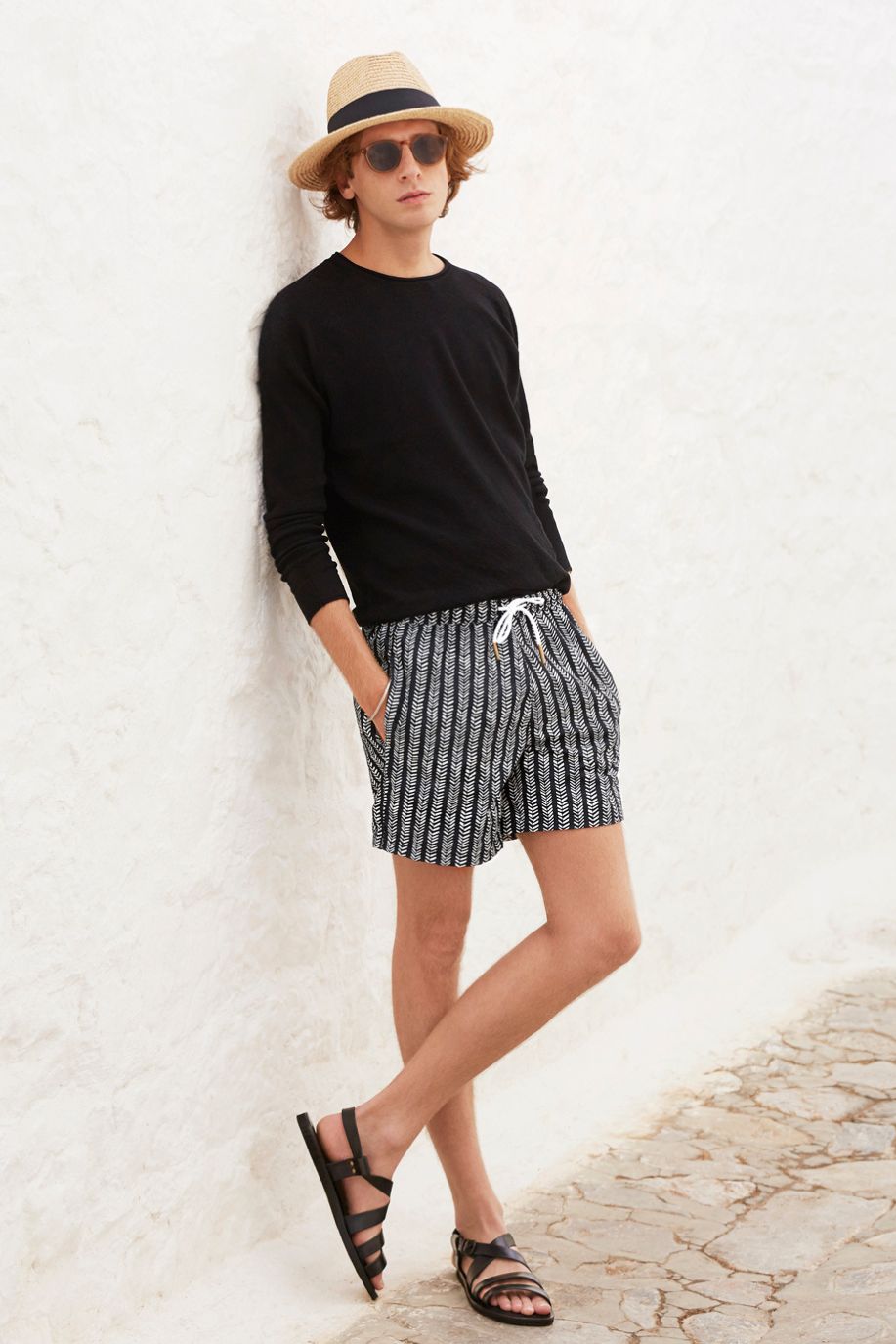 A man in patterned Sea You Soon swim shorts, a black top, sandals, and a straw hat leans against a white stone wall on a sunlit Mediterranean street.