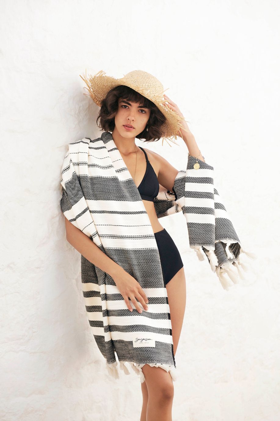 A woman in a black bikini and straw hat drapes a striped Sea You Soon Tencel beach towel over her shoulders while standing against a bright white Mediterranean wall.