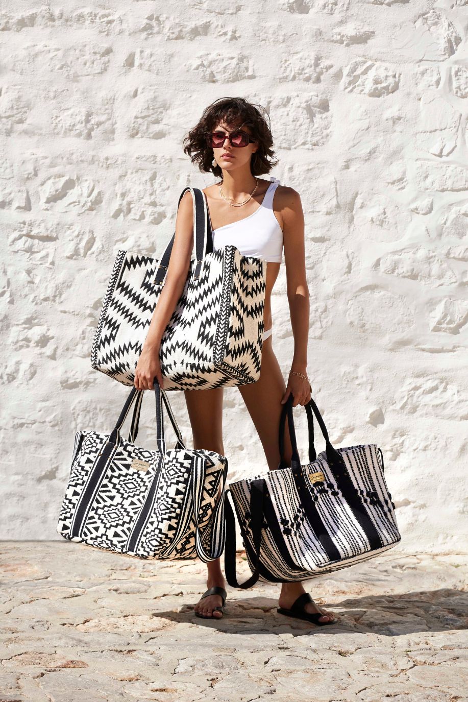 A woman in a white bikini top holds three Sea You Soon oversized beach tote bags with different black-and-white woven patterns, standing against a bright stone wall.