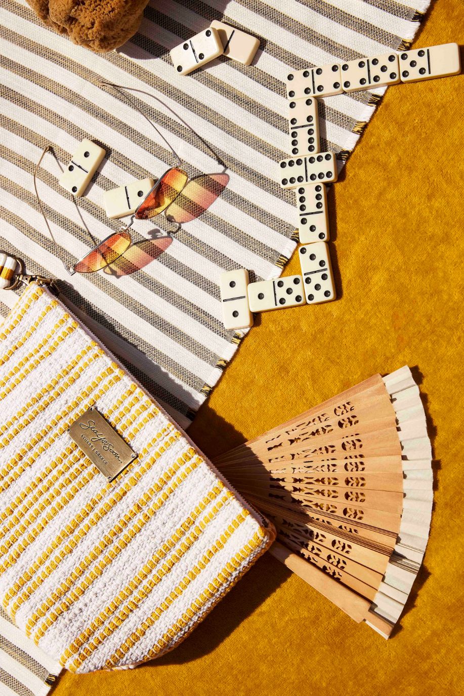 A flatlay of a Sea You Soon woven clutch, striped beach towel, sunglasses, domino pieces and a wooden hand fan styled on warm yellow fabric.