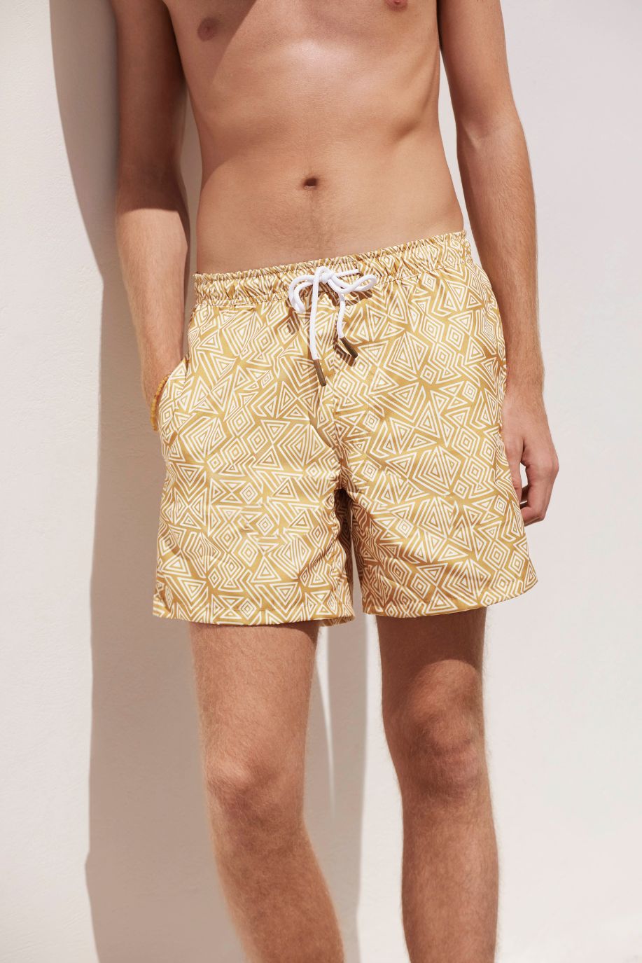 A man stands against a sunlit wall wearing mustard patterned Sea You Soon swim shorts, with his hands in his pockets and soft shadows falling across the scene.