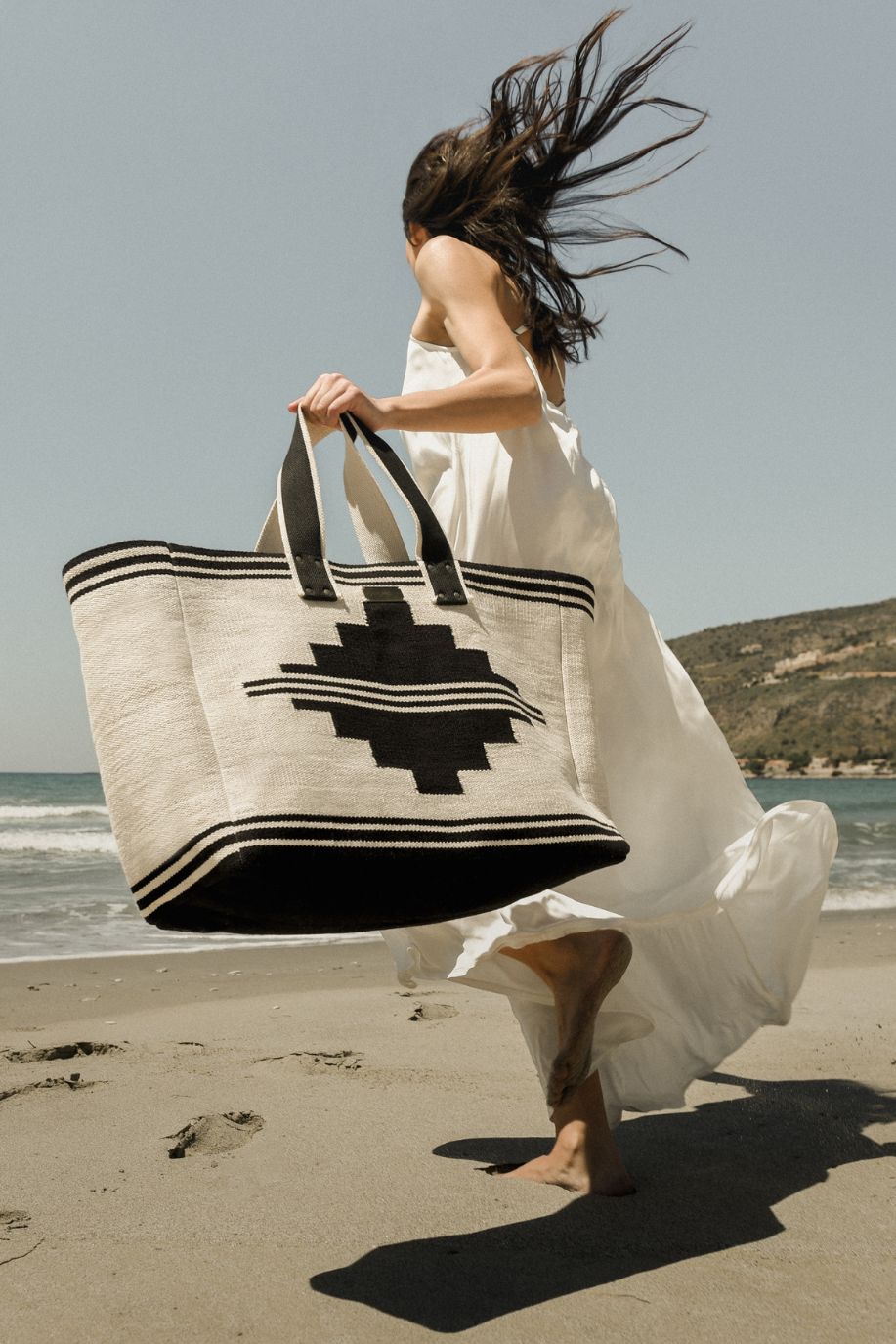 A woman runs barefoot along the beach carrying a large black-and-white Sea You Soon woven tote, her white dress moving with the coastal wind.