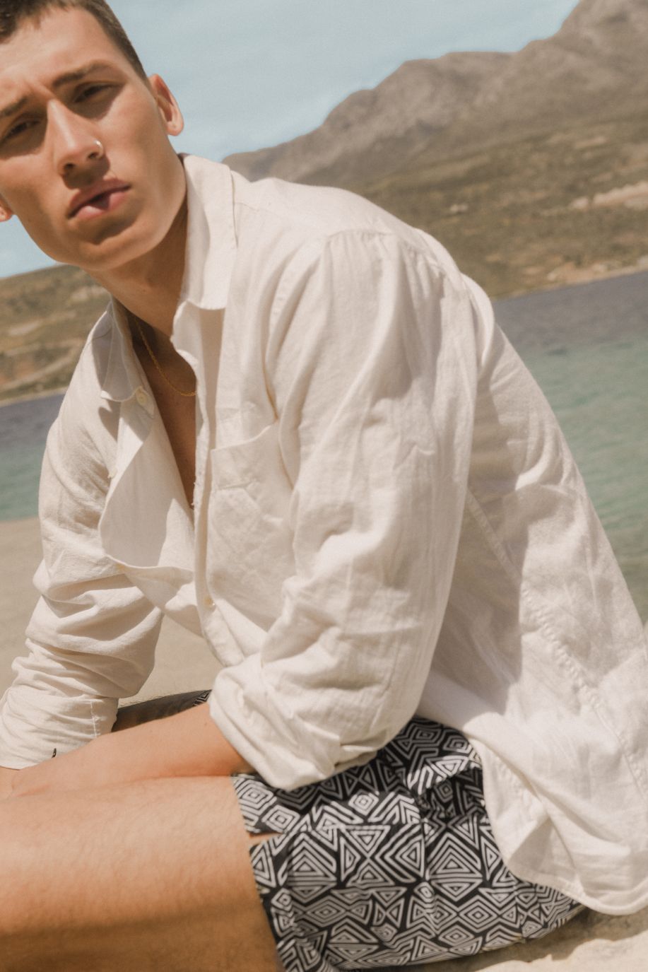 A man sits by the shoreline in a relaxed linen shirt and Sea You Soon geometric swim shorts, with rocky hills rising behind him.