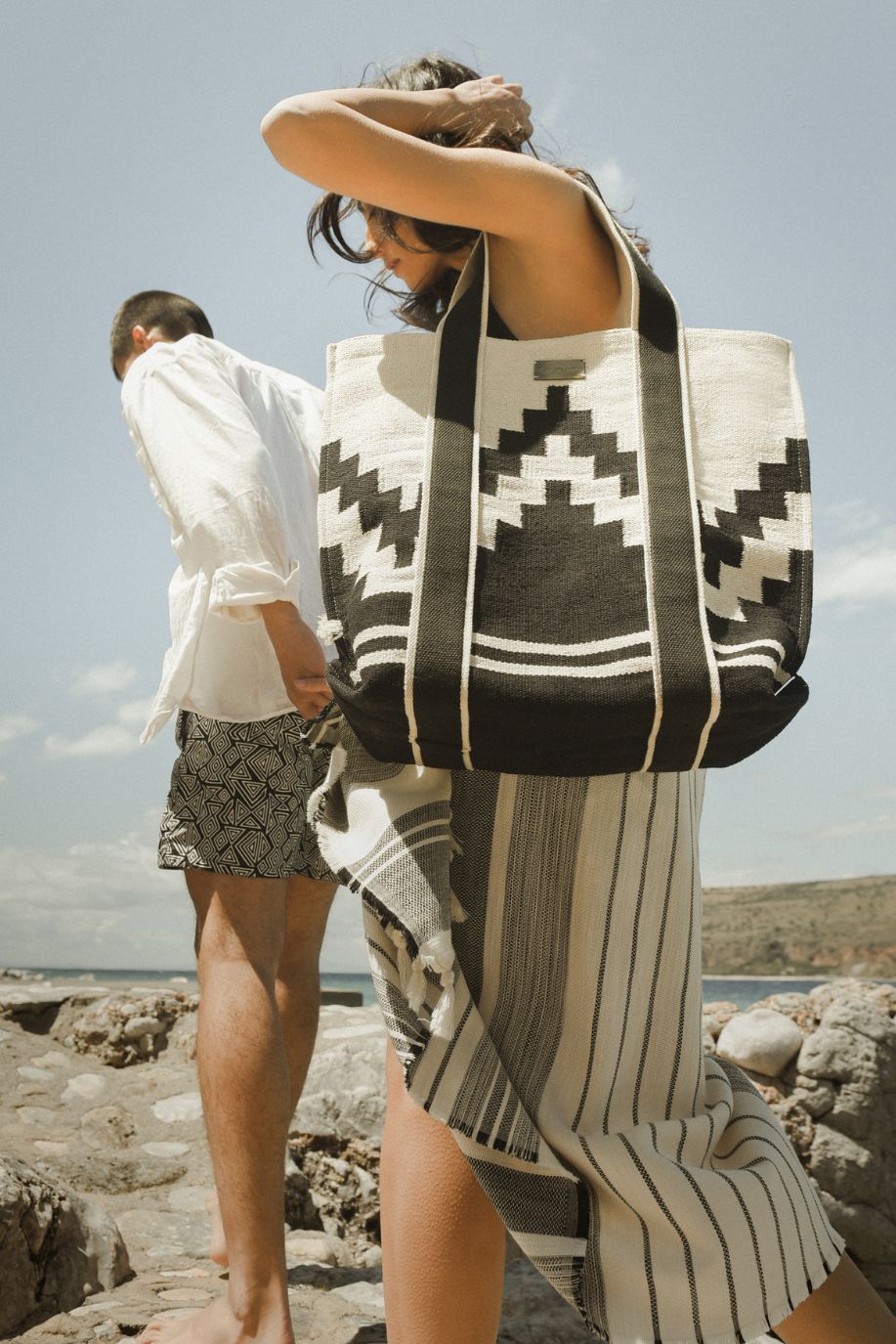 A woman walks the rocky coast carrying a black-and-white Sea You Soon woven tote bag and a striped Tencel beach towel beside a man in geometric patterned swim shorts.