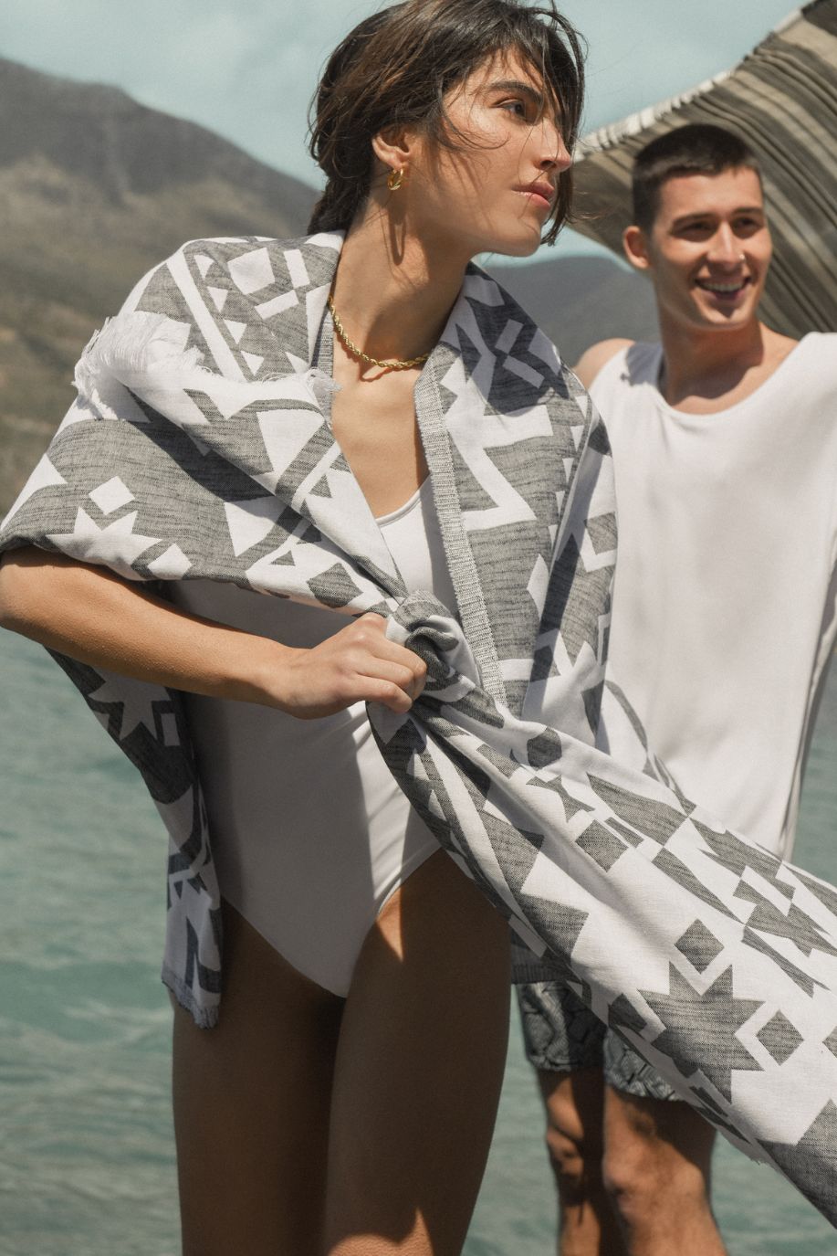 A woman stands by the sea wrapped in a grey-and-white Sea You Soon patterned beach towel as a man in patterned swim shorts smiles behind her.