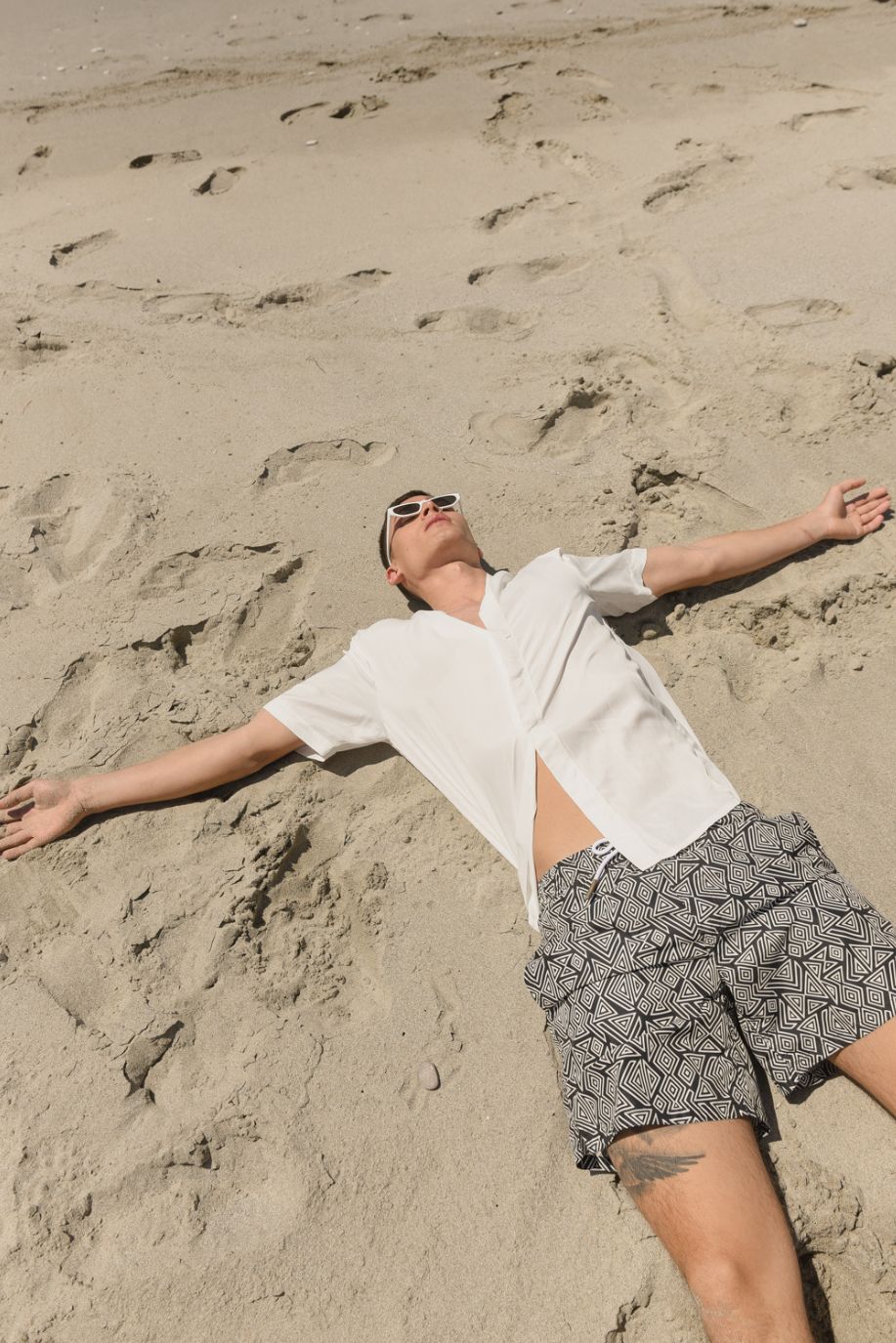 A man lies on warm sand wearing a white shirt and Sea You Soon patterned swim shorts, surrounded by footprints along the quiet shore.