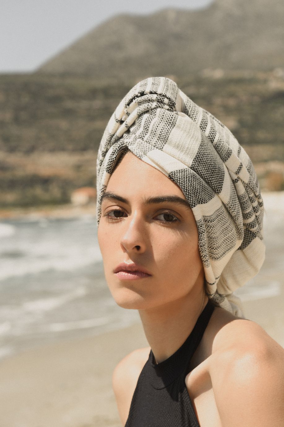 A woman gazes toward the shoreline with a striped Sea You Soon Tencel beach towel wrapped as a headscarf, waves and mountains in the distance.