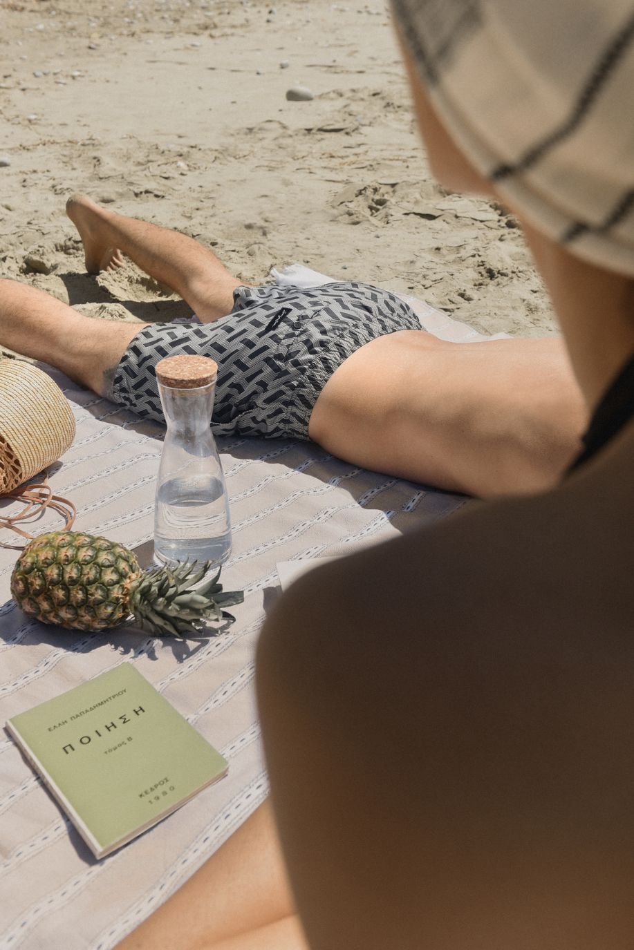 A man rests on a beach blanket in Sea You Soon geometric swim shorts beside a carafe, pineapple, and book, viewed from a woman in a towel turban.