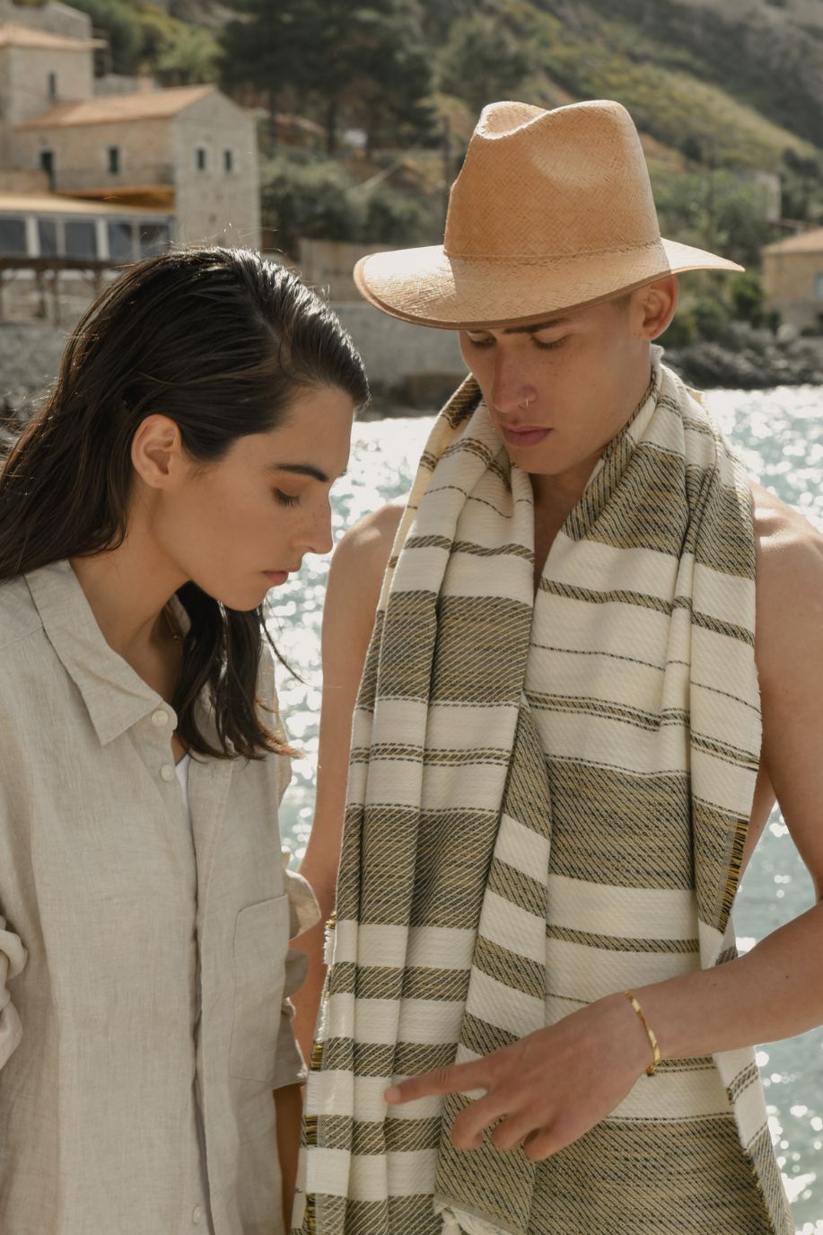 A man drapes a striped Sea You Soon Tencel beach towel over his shoulders beside a woman in a linen shirt as they overlook the calm, sunlit waterfront.