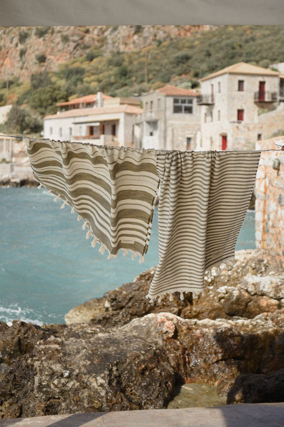Two striped Sea You Soon Tencel beach towels hang on a line above rocky seaside cliffs, moving gently in the breeze with stone houses rising behind them.