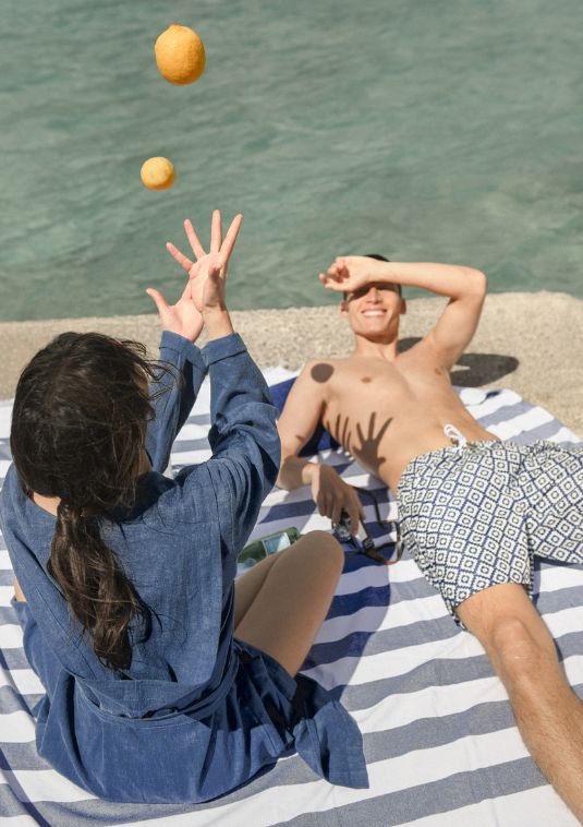 Two people relaxing by the water on a striped beach blanket, one wearing Sea You Soon patterned swim shorts while tossing lemons in soft coastal light.