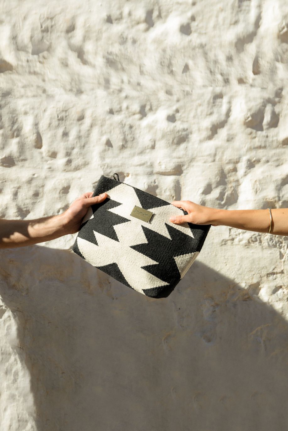 Two hands exchange a small black-and-white patterned woven Sea You Soon clutch in bright sunlight against a textured white wall, capturing a simple Mediterranean moment.