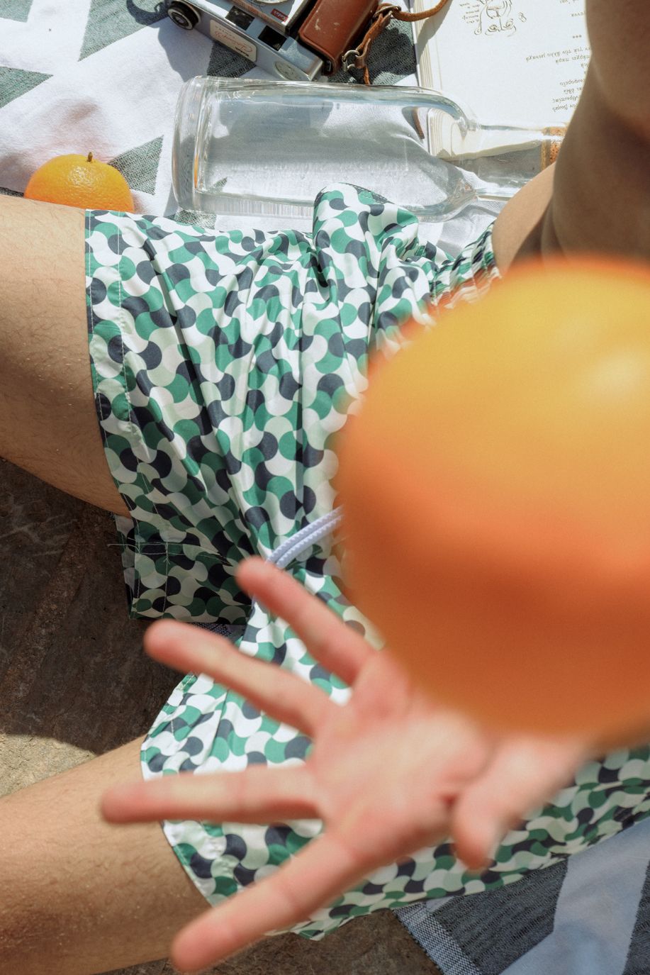 Close-up of a man in emerald geometric patterned Sea You Soon swim shorts reaching for a tossed orange, surrounded by summer essentials during a relaxed Mediterranean moment.