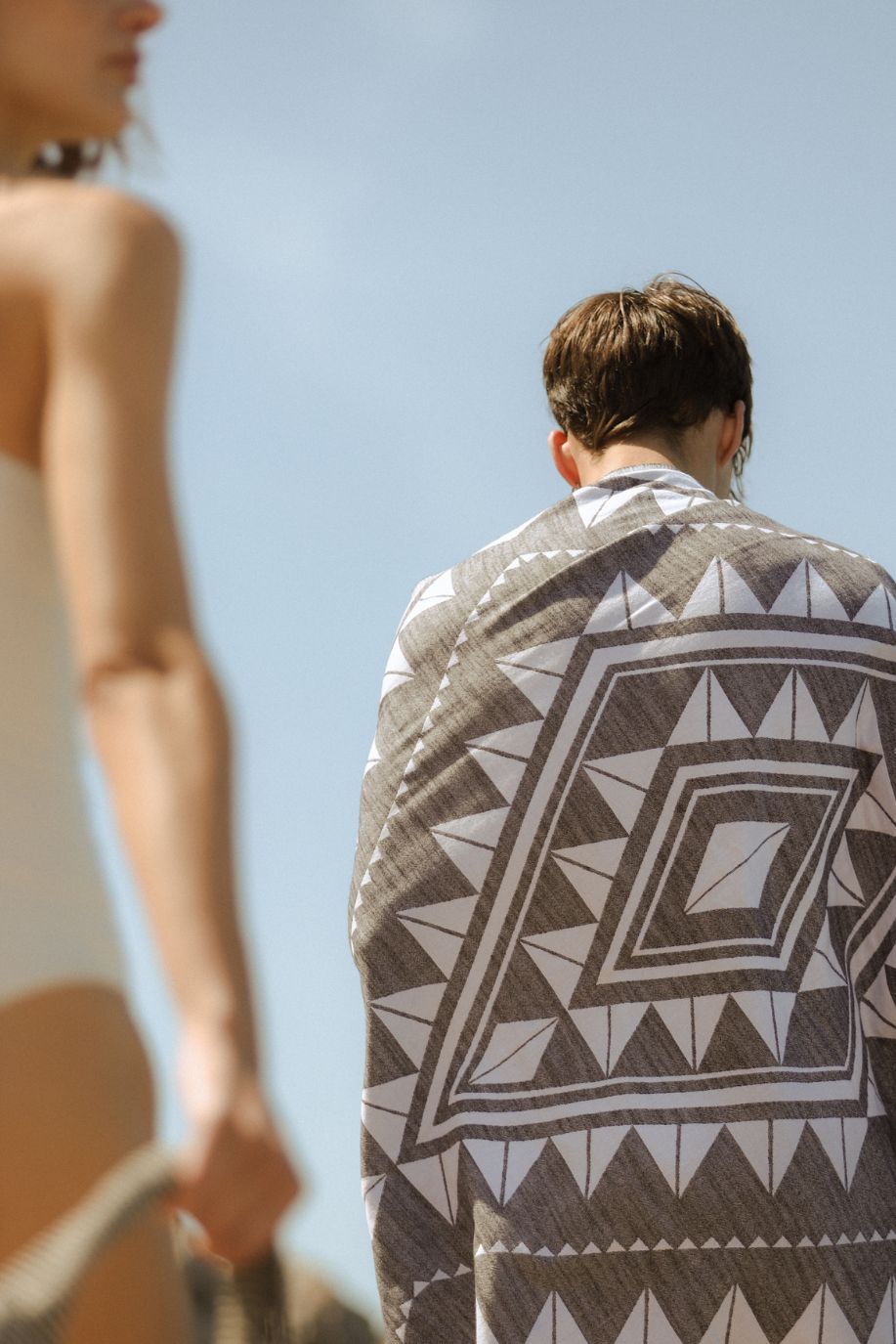 Man stands with his back to the camera wrapped in a large geometric patterned Sea You Soon beach towel, while a woman in a white swimsuit walks past under a clear sky.