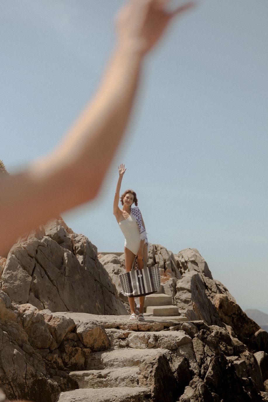 Woman in a white swimsuit waves from rocky seaside steps, holding a black-and-white striped woven Sea You Soon tote bag and a patterned towel against a soft Mediterranean backdrop.
