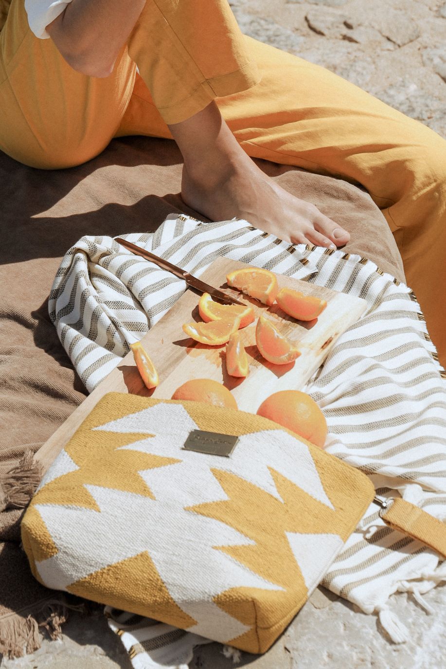 Sliced oranges rest on a wooden board beside Lightweight striped beach towels crafted from Tencel and a yellow geometric patterned Sea You Soon woven clutch, with a person in yellow trousers lounging in a relaxed beach setting.