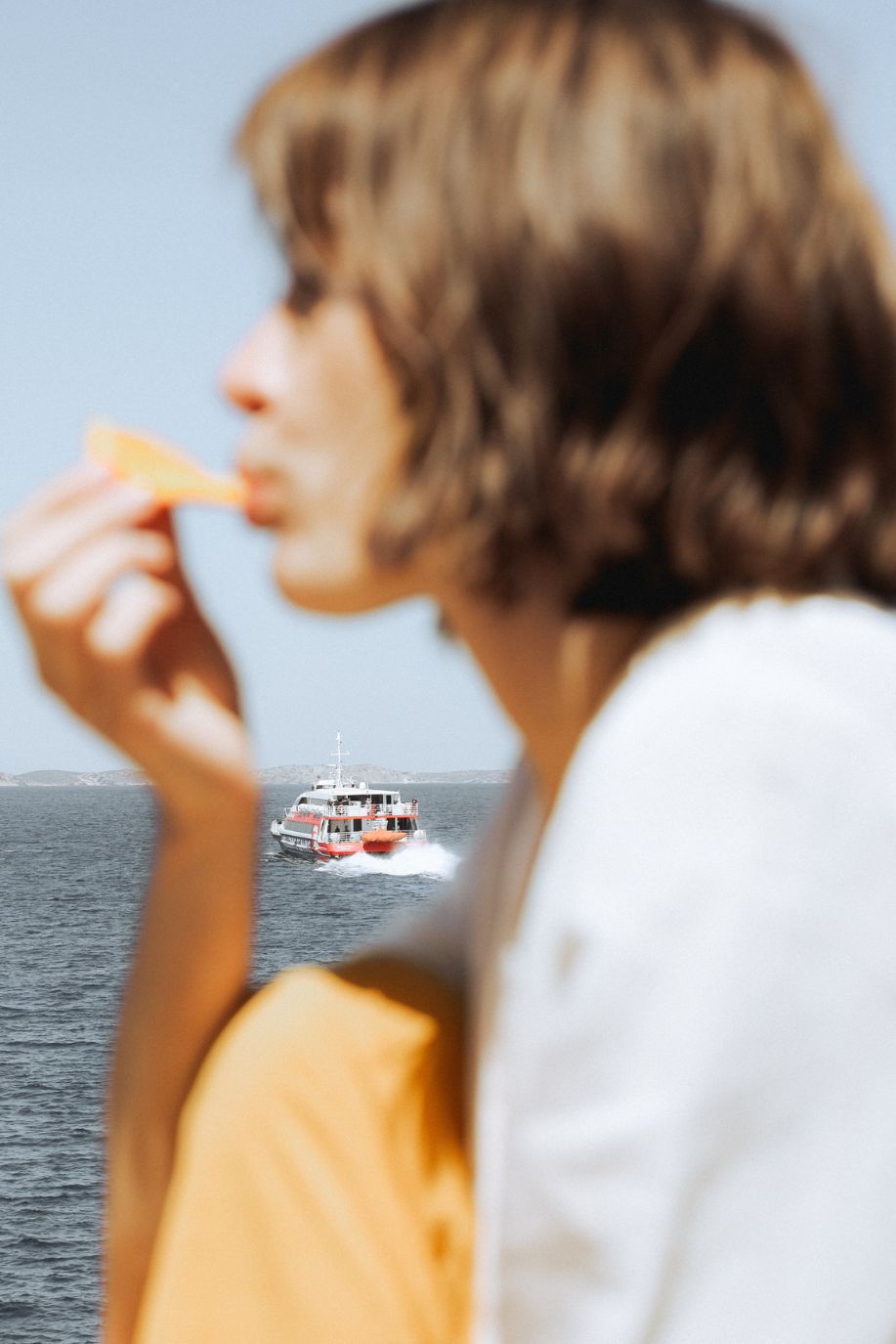 Woman eating an orange slice by the sea as a small ferry passes in the distance, creating a soft, sunlit Mediterranean moment with a relaxed summer feel.