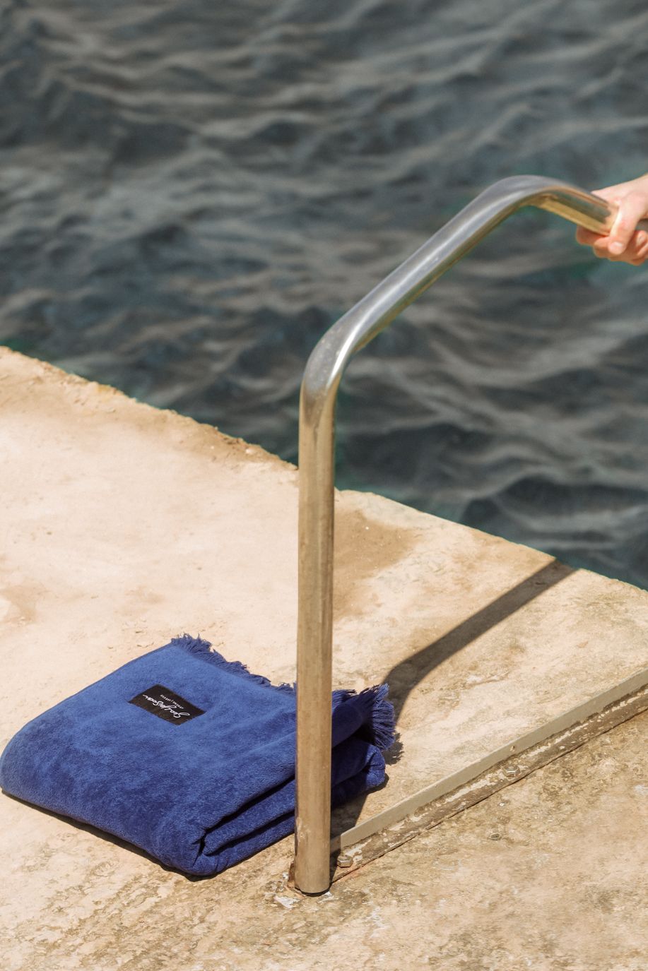 A folded deep blue fringed Sea You Soon cotton velour beach towel rests on a stone platform beside a metal ladder, overlooking gently moving water on a quiet coastal afternoon.