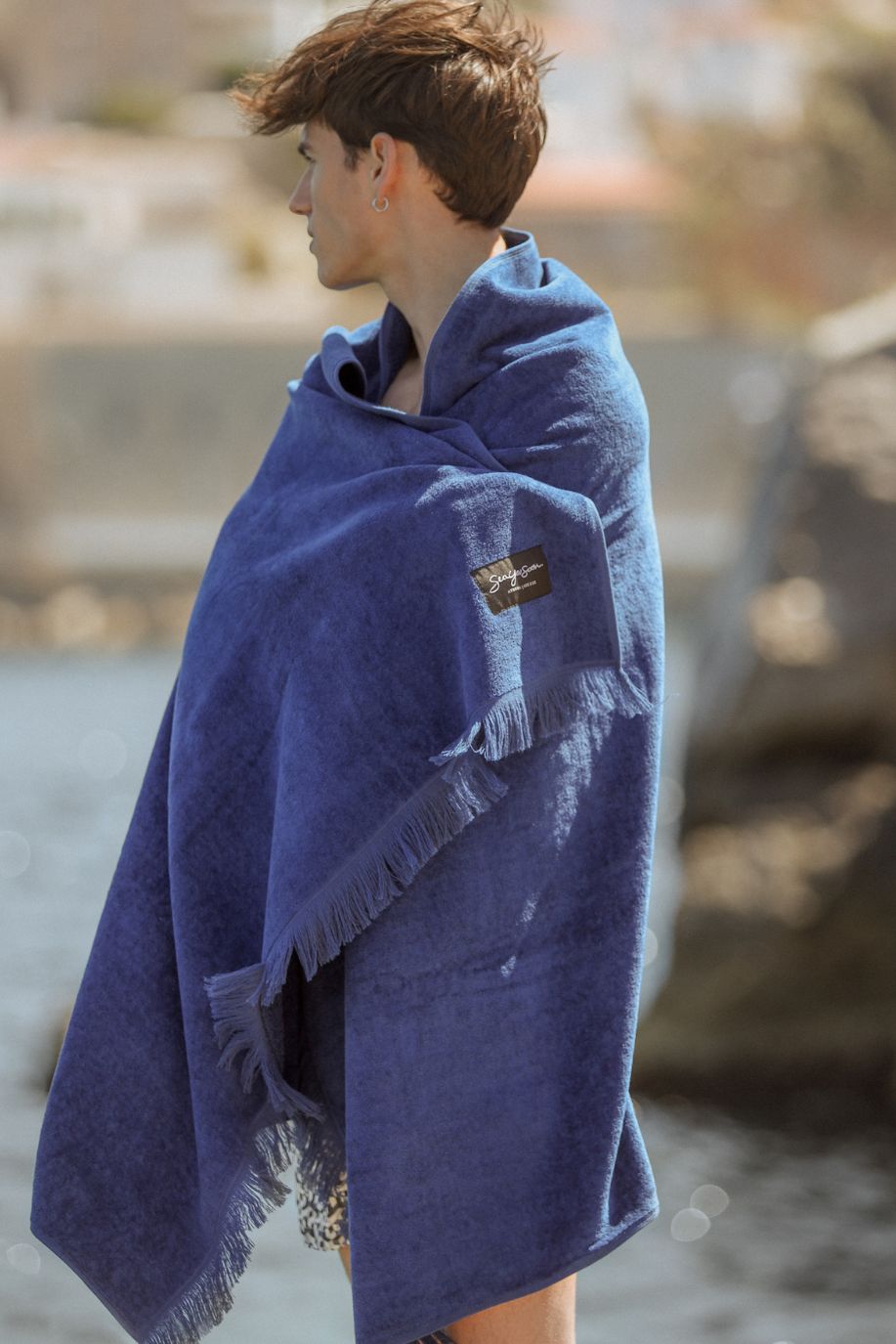 A man stands by the water wrapped in a deep blue, fringed Sea You Soon velour cotton beach towel, looking toward the shoreline in a calm Mediterranean setting with soft coastal light.