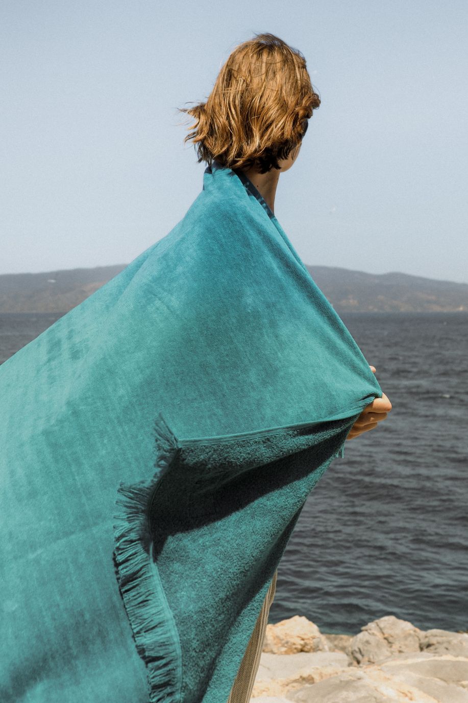 Woman facing the sea with a large emerald fringed Sea You Soon velour cotton beach towel draped around her shoulders, standing on rocky shorelines under bright Mediterranean sky.