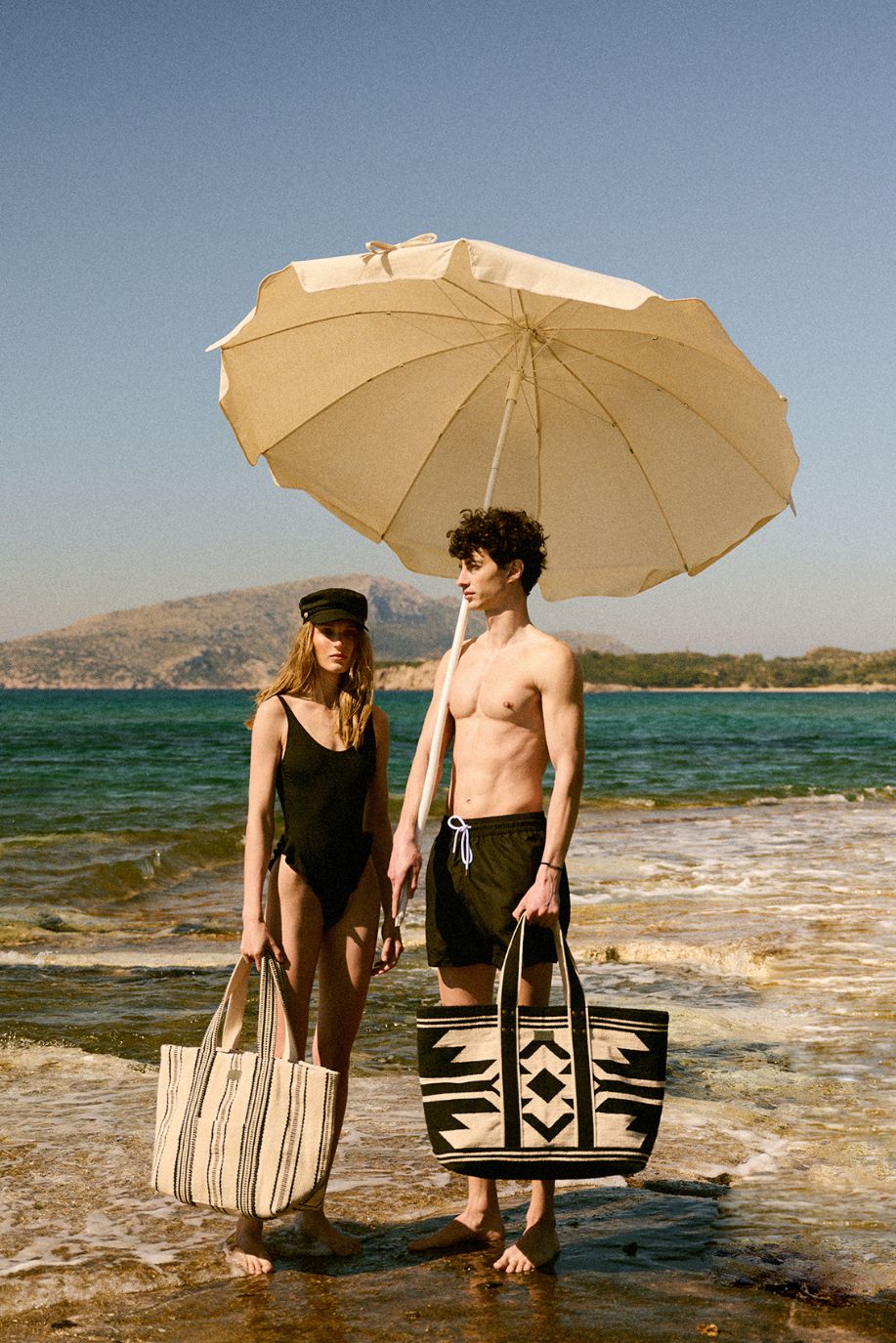 A man and woman stand by the shoreline under a large umbrella, each holding a woven beach tote with striped and patterned designs against the calm sea.
