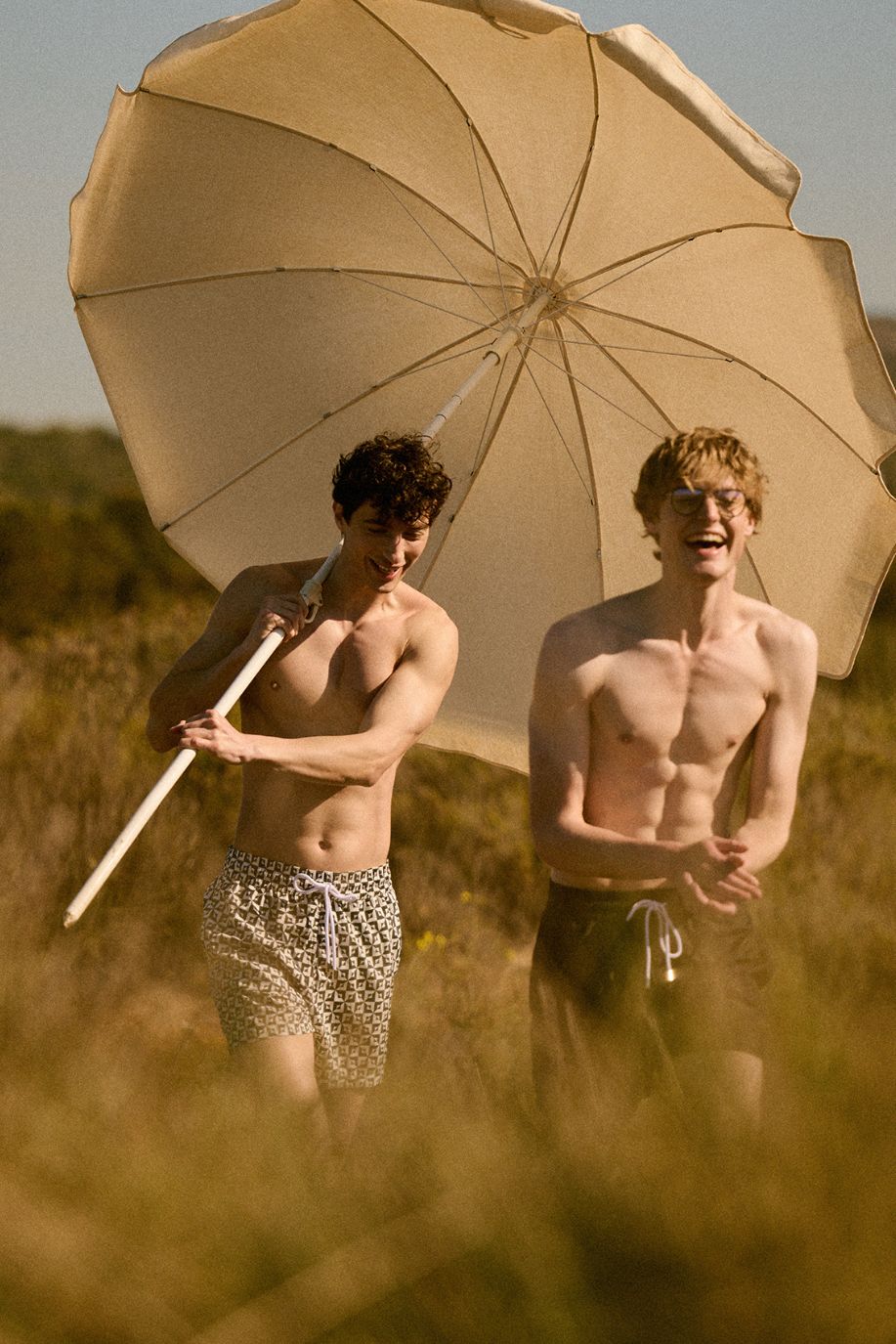 Two men walk through sunlit coastal fields, one carrying a beach umbrella, both wearing Sea You Soon patterned swim shorts in light summer tones.