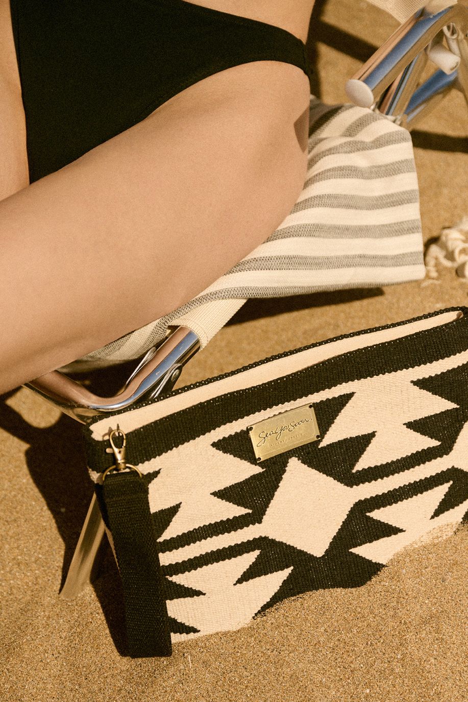 A woman sits on a sandy beach lounger beside a black-and-white patterned, woven Sea You Soon clutch resting against a striped Tencel beach towel and sunlit metal.