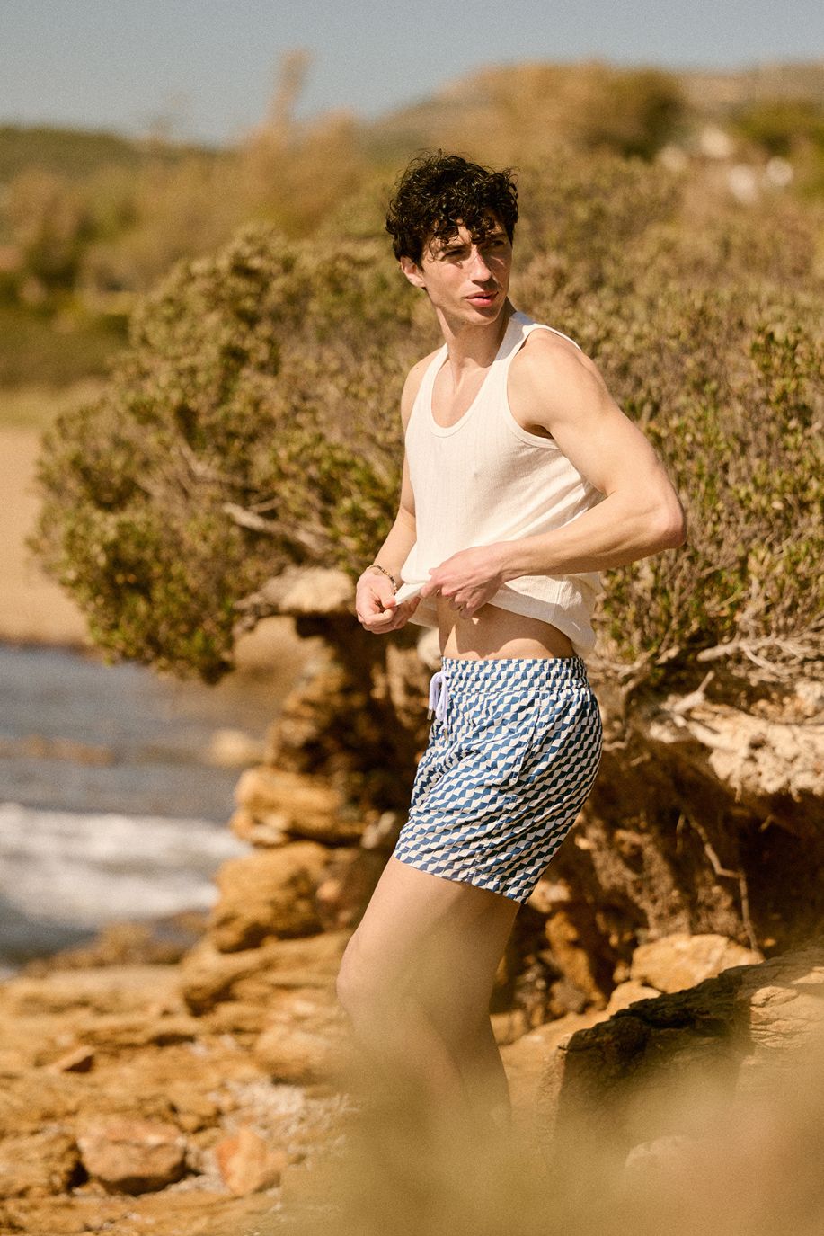 A man stands near the rocky coast in the sun, wearing blue patterned Sea You Soon swim shorts paired with a casual white tank top.