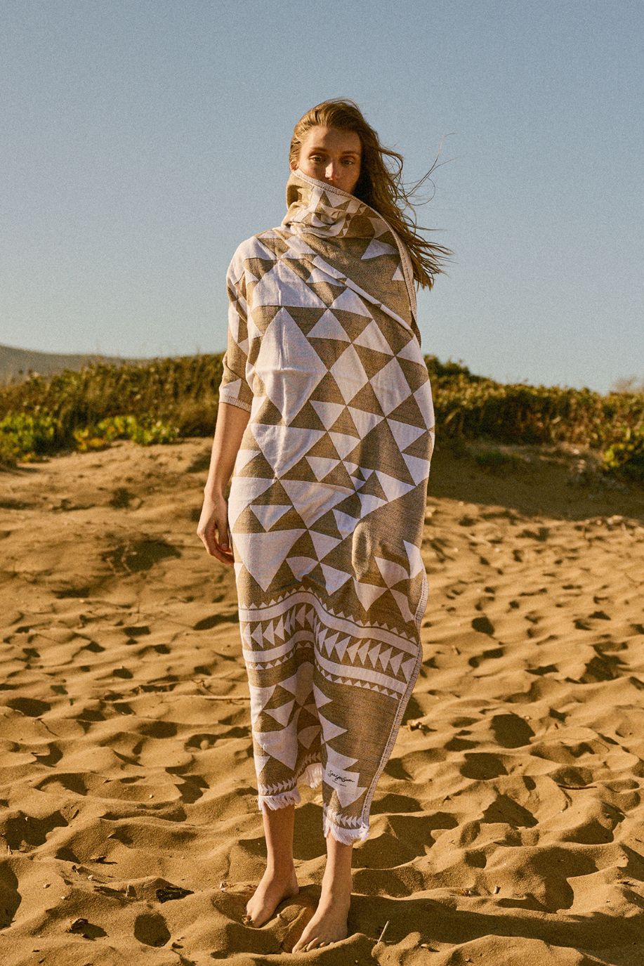 A woman stands wrapped in a geometric lightweight beach towel on a sunlit sandy shore.