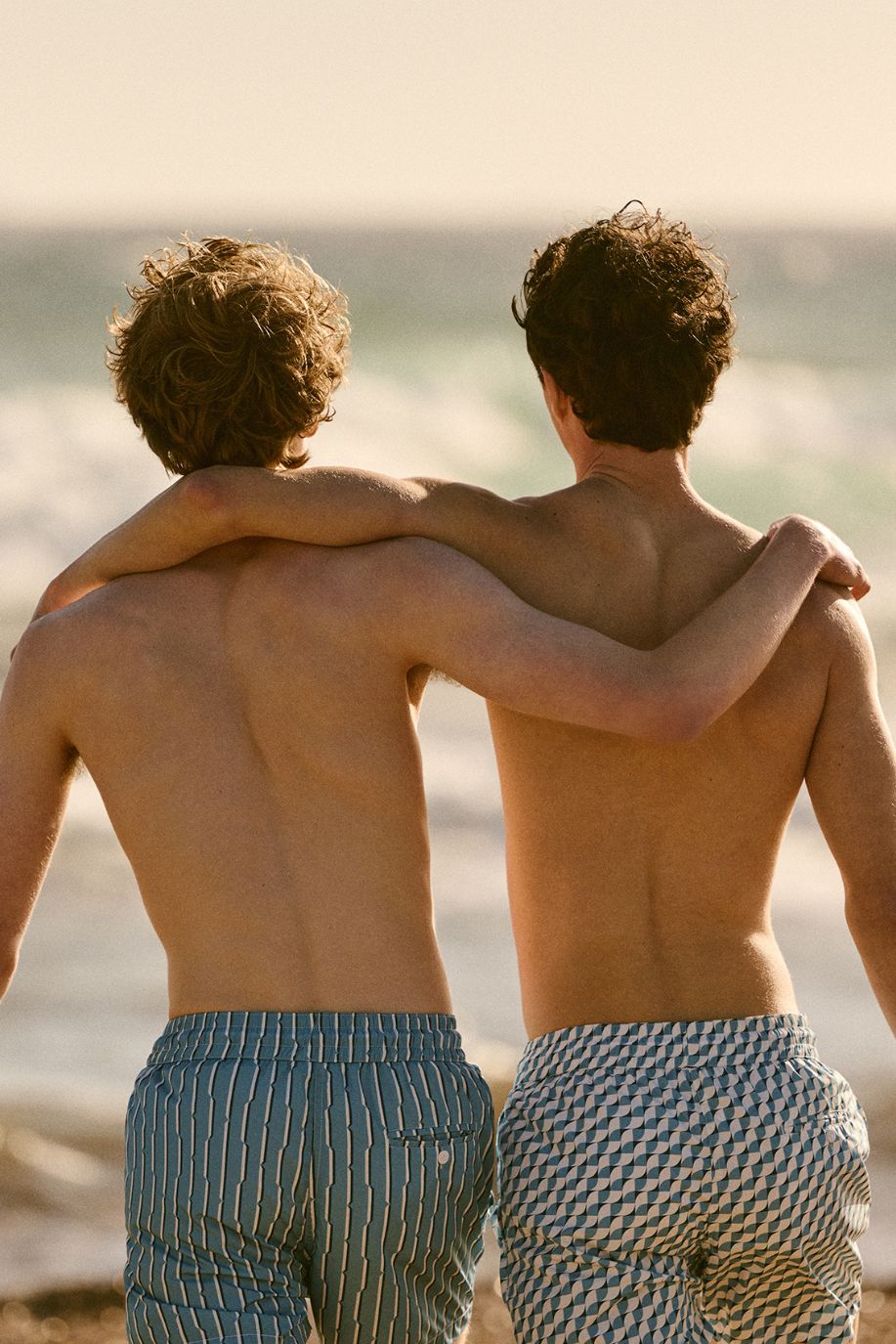 Two men walk along the shoreline in patterned swim shorts, their arms around each other as the waves roll in behind them under warm evening light