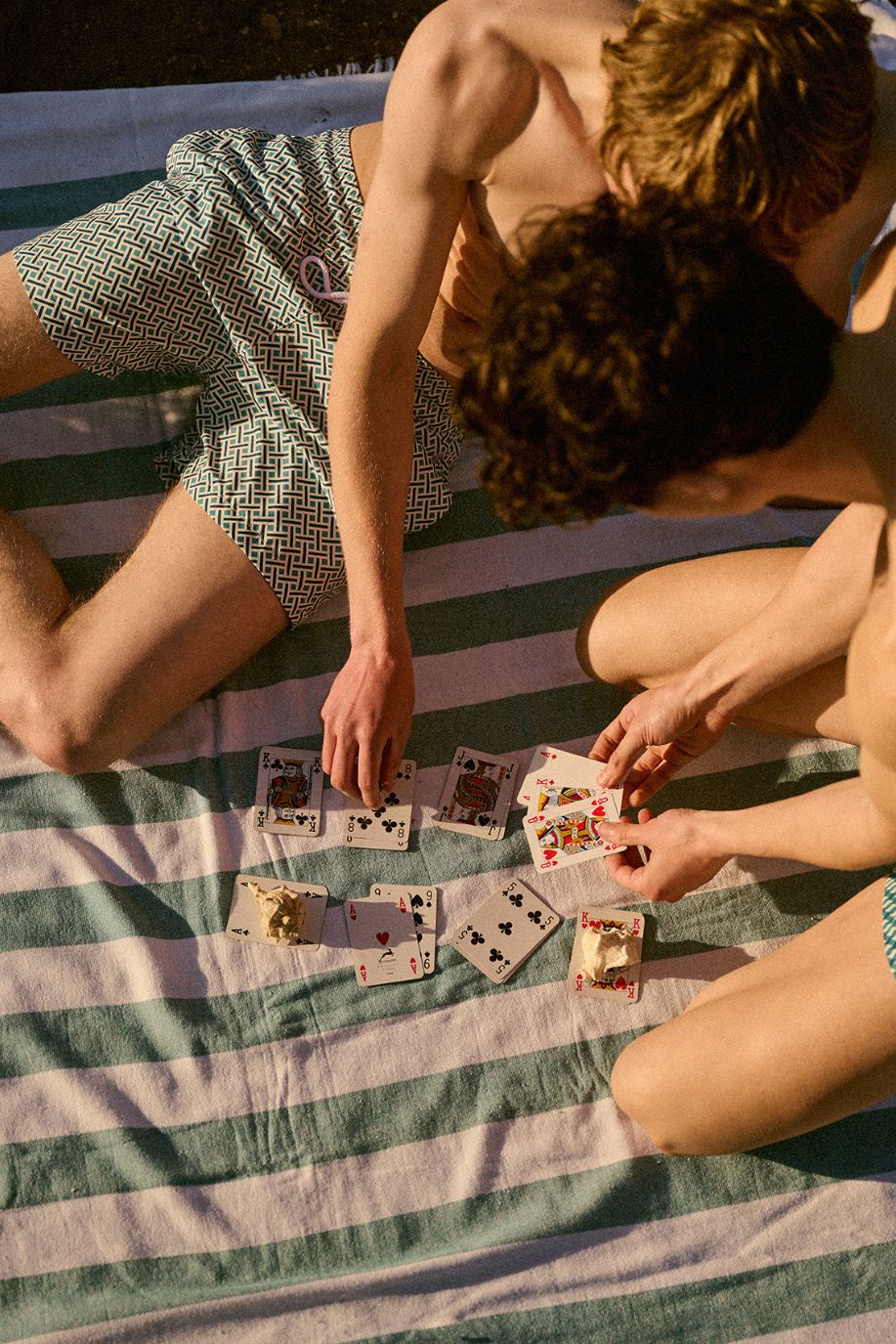 Two men sit on a striped beach blanket playing cards, both wearing patterned swim shorts with the late sun casting soft shadows across the sand.