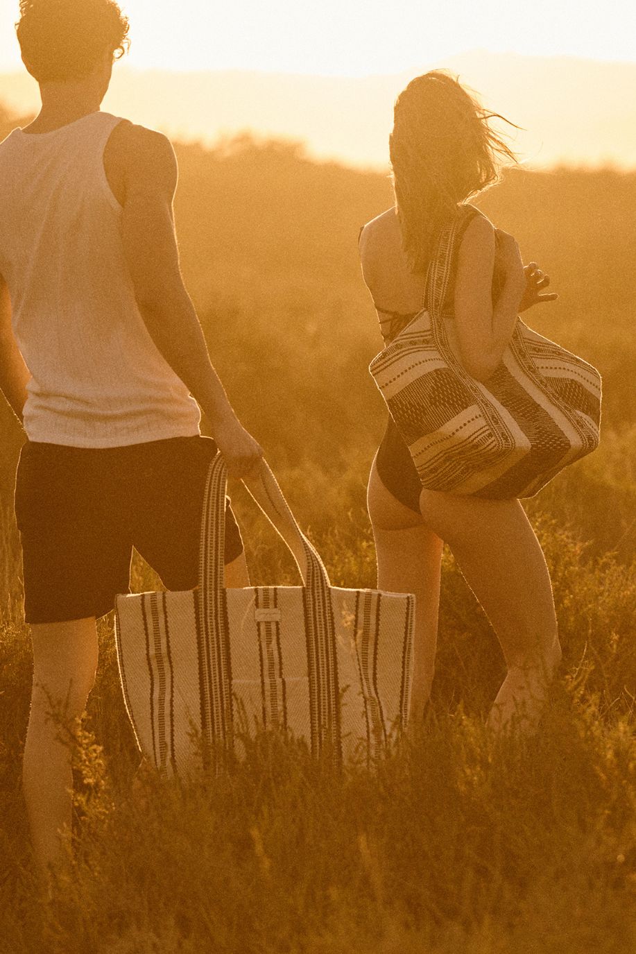 A man and woman walk through golden dusk carrying woven beach tote bags, their swimwear and the warm light creating a soft summer silhouette.