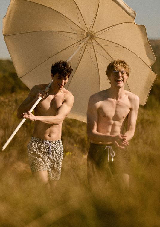 Two men wearing Sea You Soon patterned swim shorts walk through tall grass under a sun umbrella, captured in warm coastal light during a relaxed summer moment.