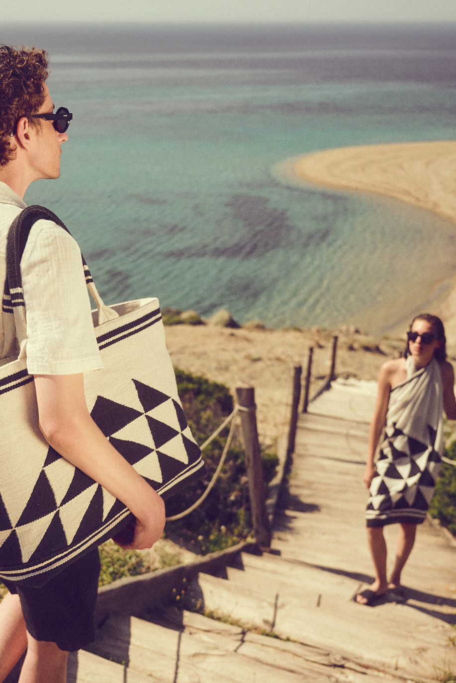 A man walks down a wooden path toward the sea carrying a geometric Sea You Soon woven tote bag, while a woman wrapped in a matching pareo waits by the shore.