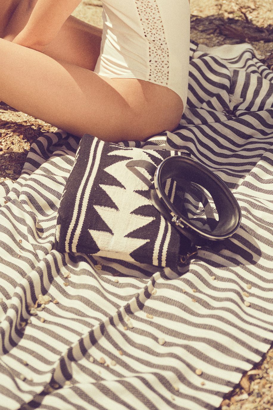 A woman in a white swimsuit sits on a striped Sea You Soon lightweight Tencel beach towel, with a woven geometric clutch and sunglasses resting in the sand beside her.