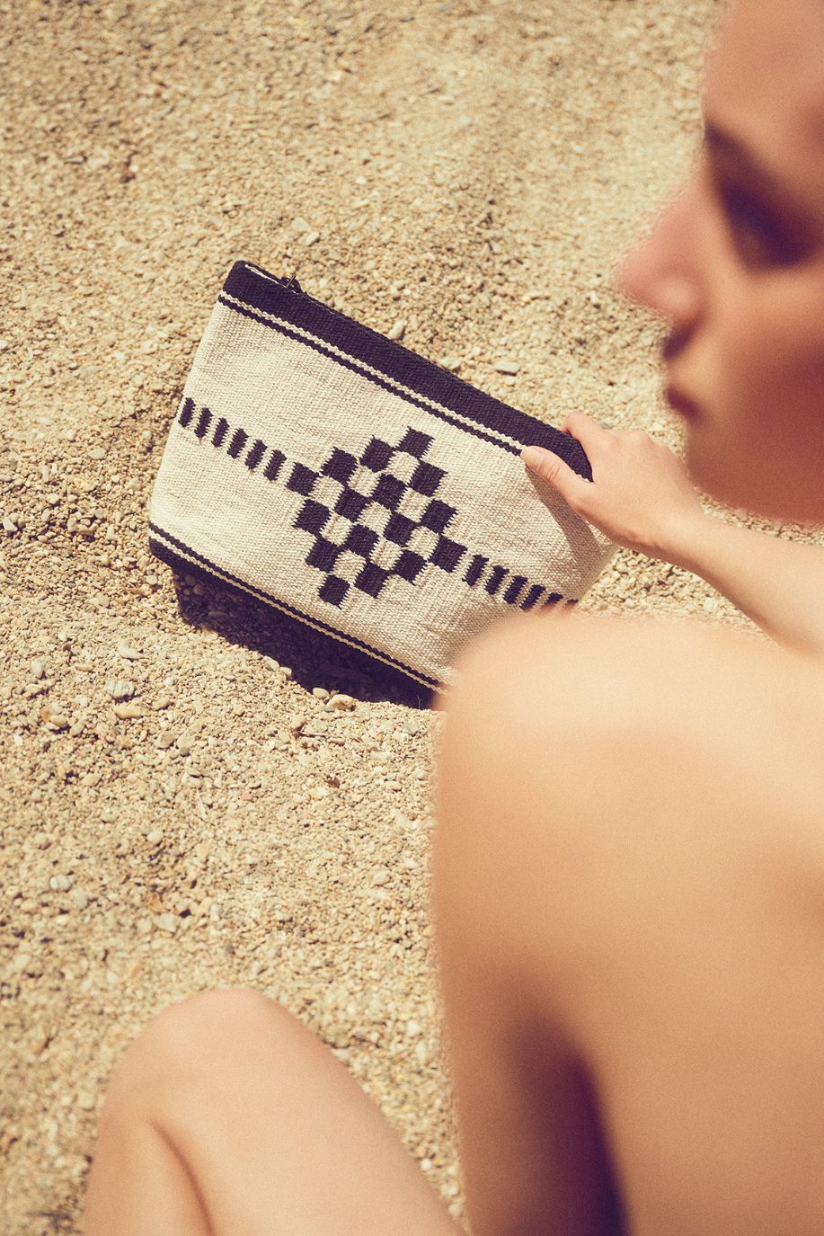 A woman sits on warm sand holding a geometric Sea You Soon pouch, its textured woven pattern contrasting with the coastal landscape around her.