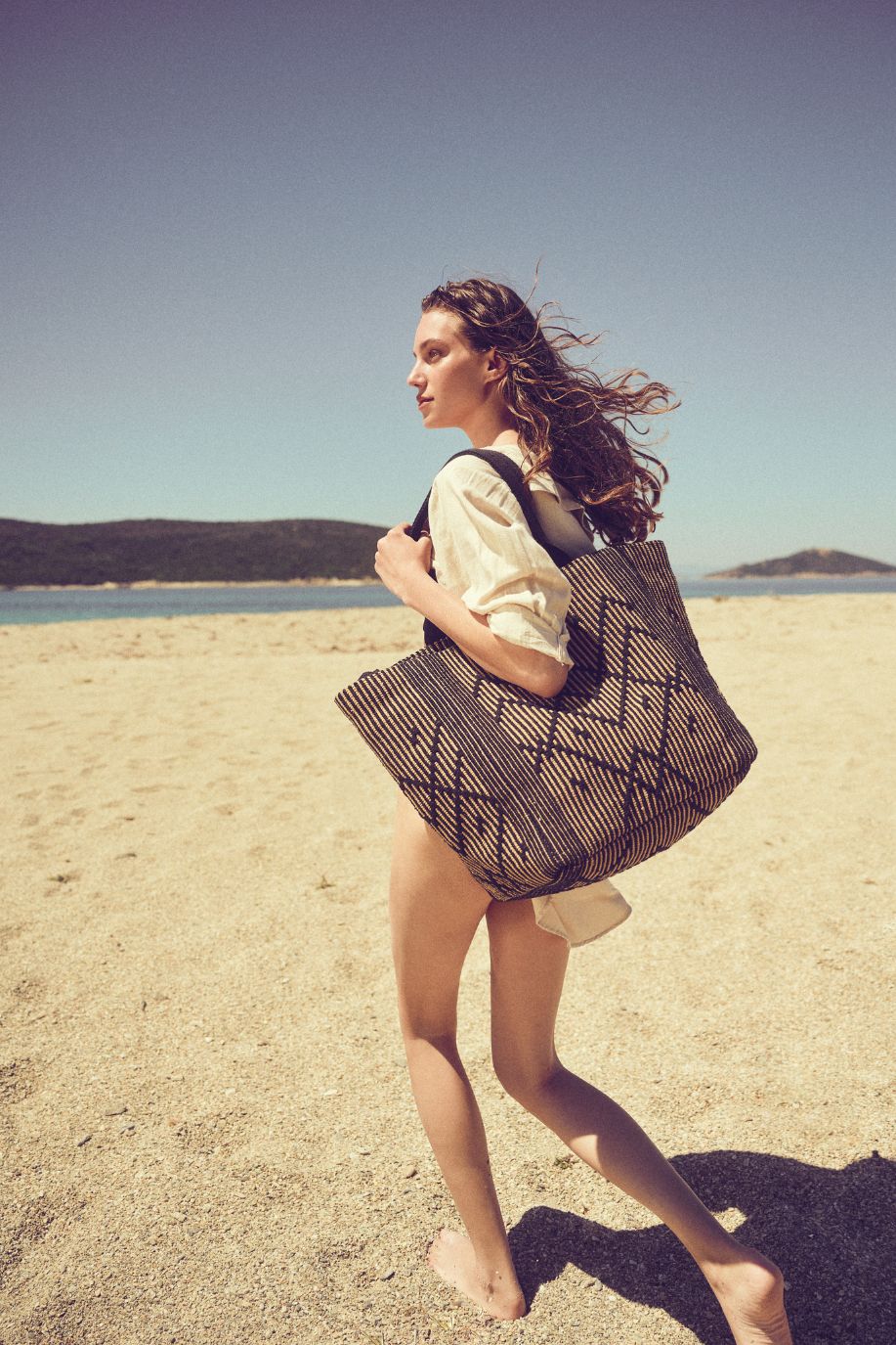 A woman walks along a sunlit Mediterranean beach carrying a woven, patterned Sea You Soon tote bag, her linen shirt moving softly in the breeze.