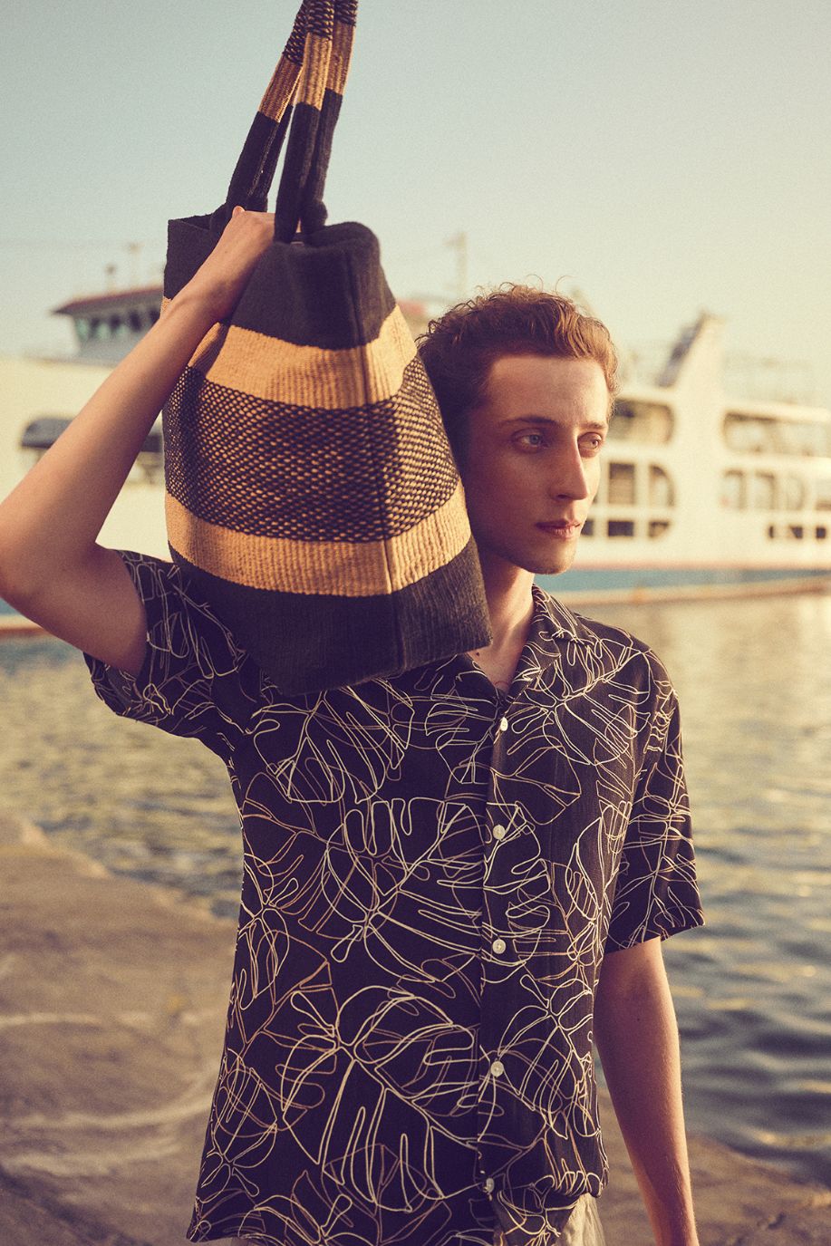 A man walks along the harbor at sunset wearing a patterned Sea You Soon resort shirt, carrying a woven striped beach tote bag over his shoulder.