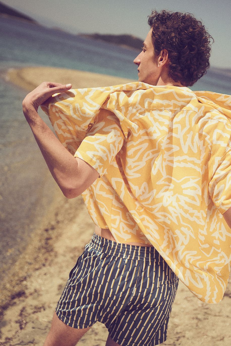A man adjusts a yellow patterned Sea You Soon resort shirt, his swim shorts catching the breeze as he walks along the shoreline on a warm day.