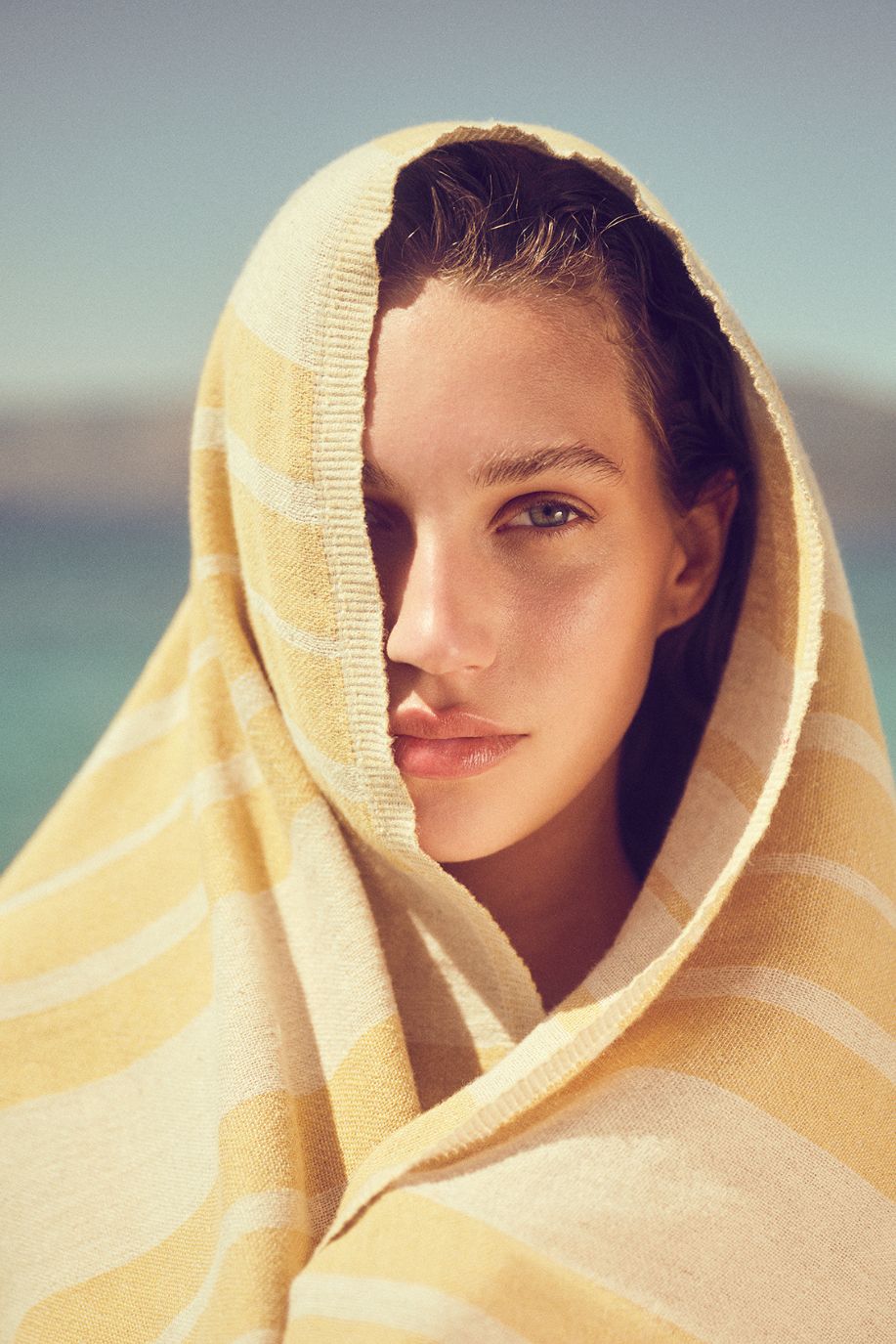 A woman wrapped in a soft yellow striped Sea You Soon linen beach towel gazes toward the camera, standing against a calm Mediterranean backdrop.