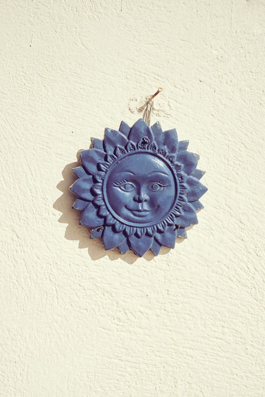A blue ceramic sun ornament hangs on a textured white wall, catching soft daylight in a simple Mediterranean setting.
